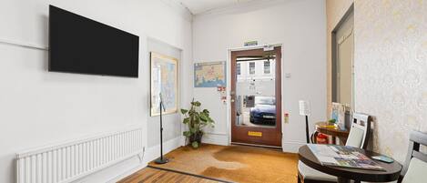 A hallway with a wooden floor, a mounted TV, a door with a sign, a potted plant, and a table with chairs.