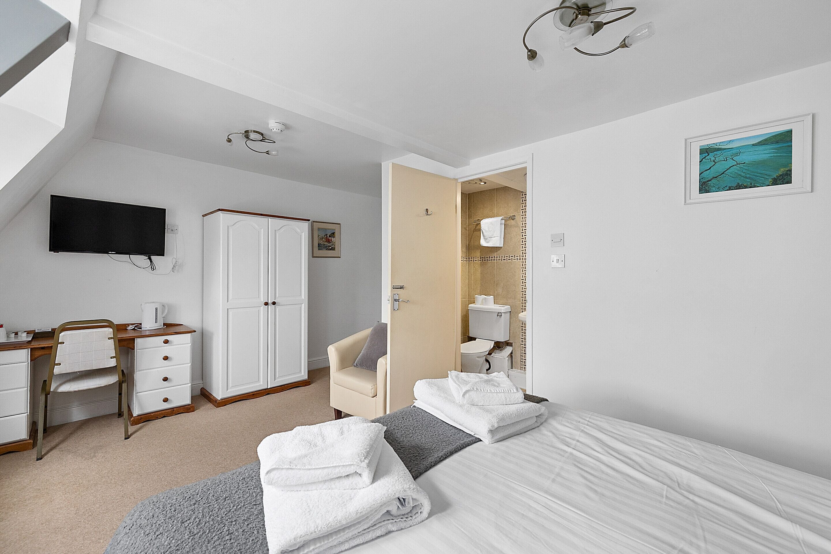 A hotel room with a bed, a desk, a chair, a TV, and a bathroom visible through an open door.