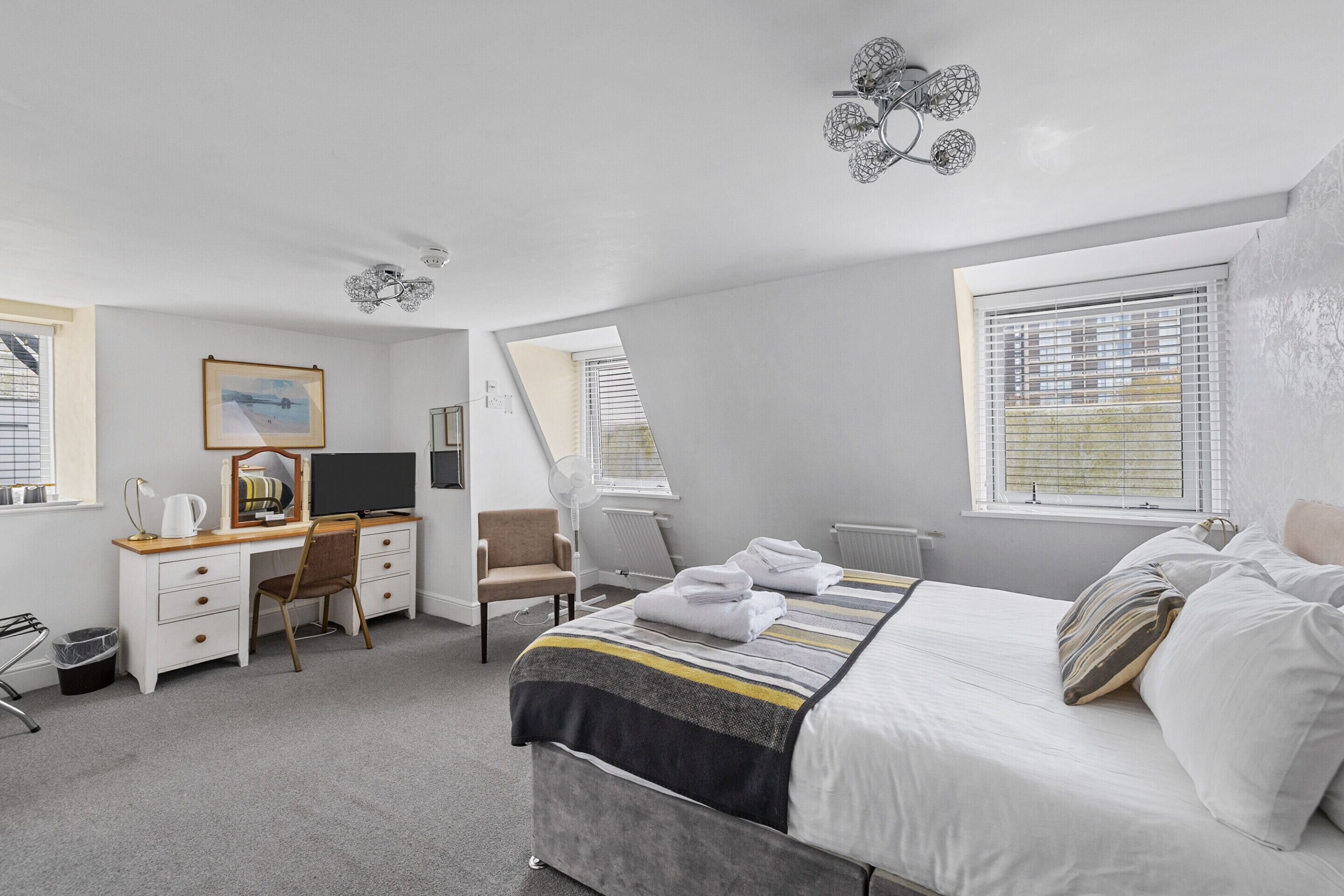 Una habitación de hotel moderna con una cama bien hecha, un escritorio con computadora, una silla y un ventanal con persianas.