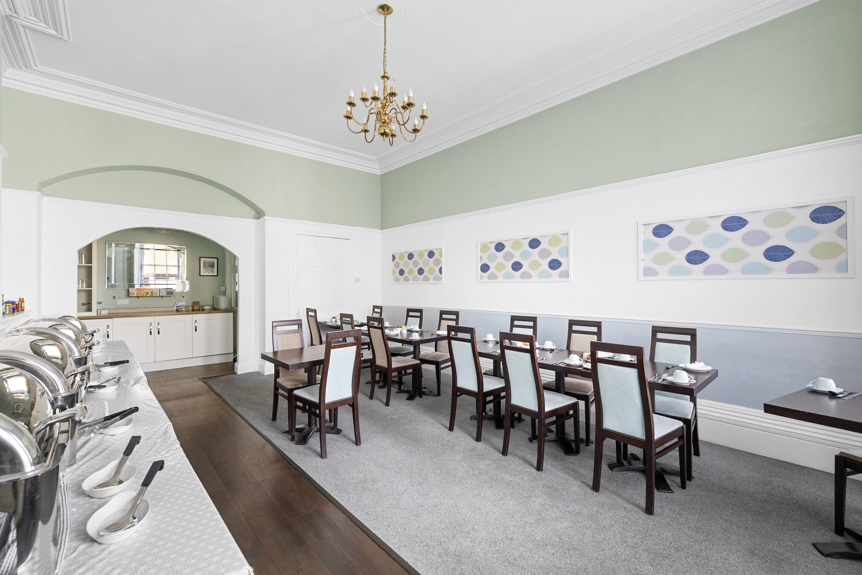 Zona pranzo con diversi tavoli e sedie, cucina sullo sfondo e decorazioni artistiche alle pareti.
