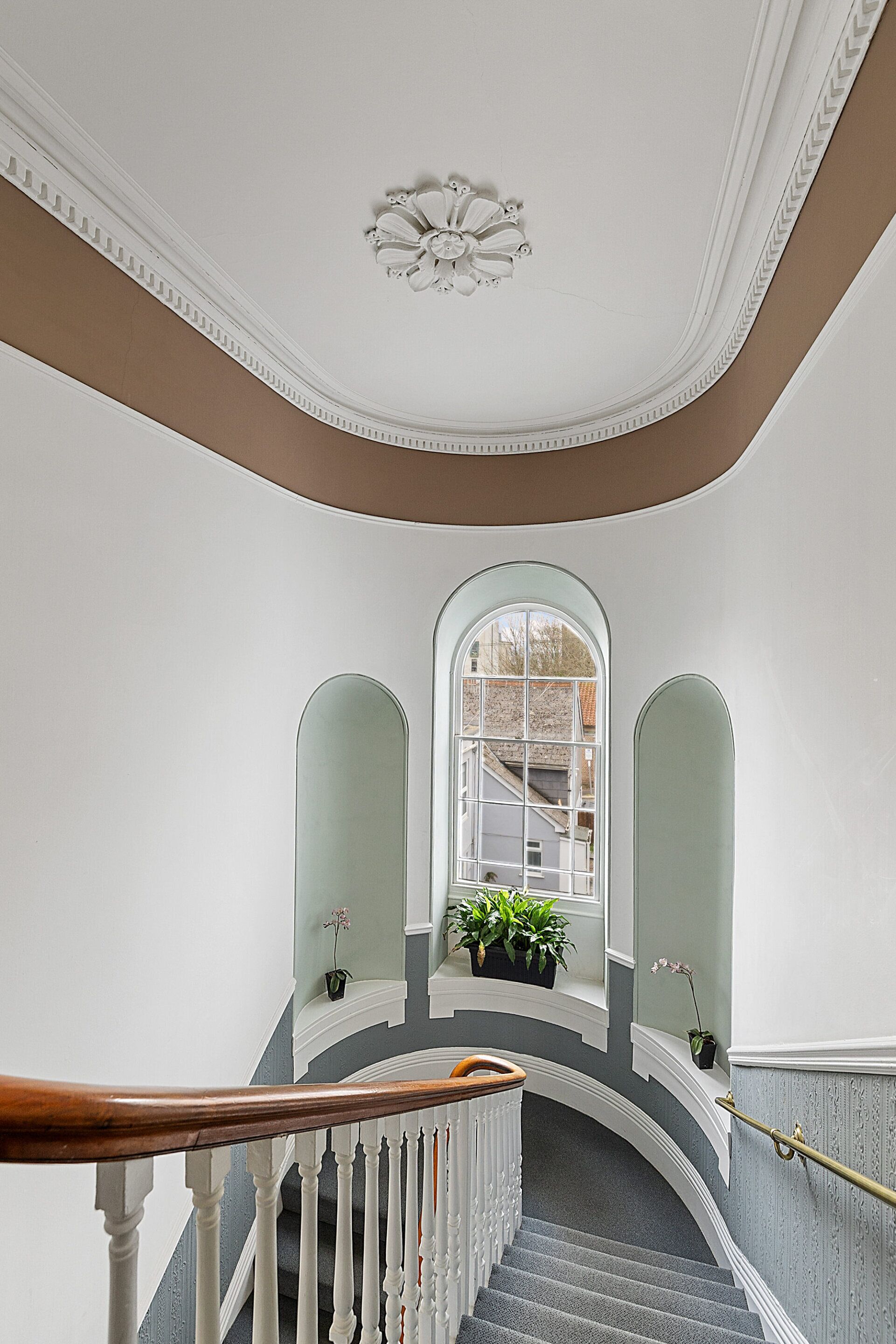 A grand staircase with white balustrades and a wooden handrail, leading to a landing with a large arched window and potted plants.