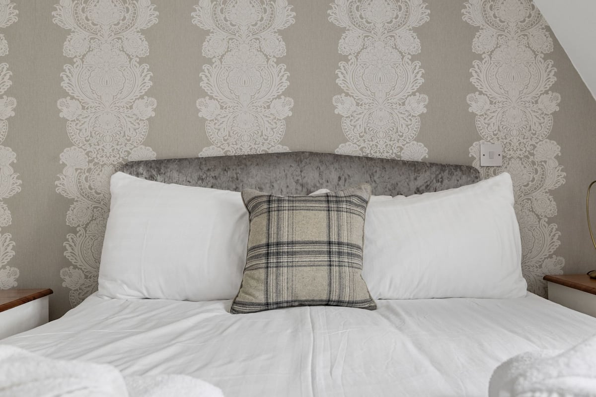 A neatly made bed with white linens, a grey headboard, and a plaid pillow.