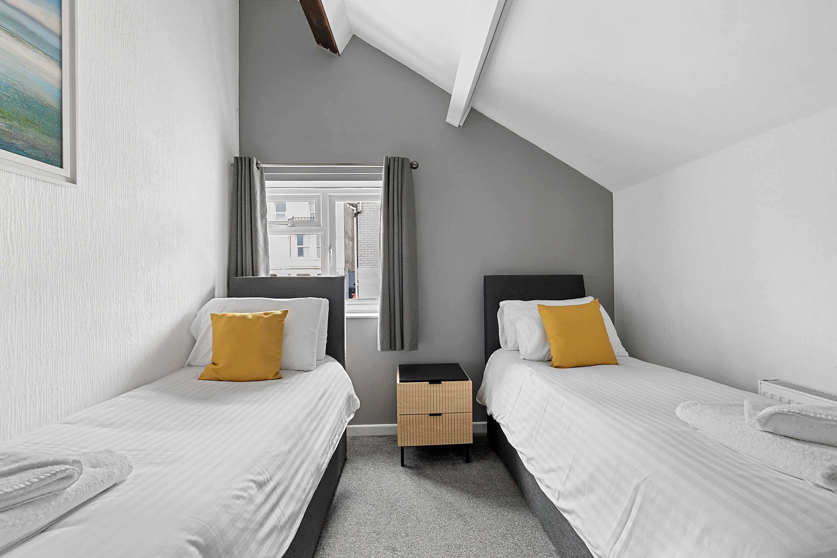 A modern hotel room with two single beds, a small wooden nightstand, a window with curtains, and a view of buildings outside.