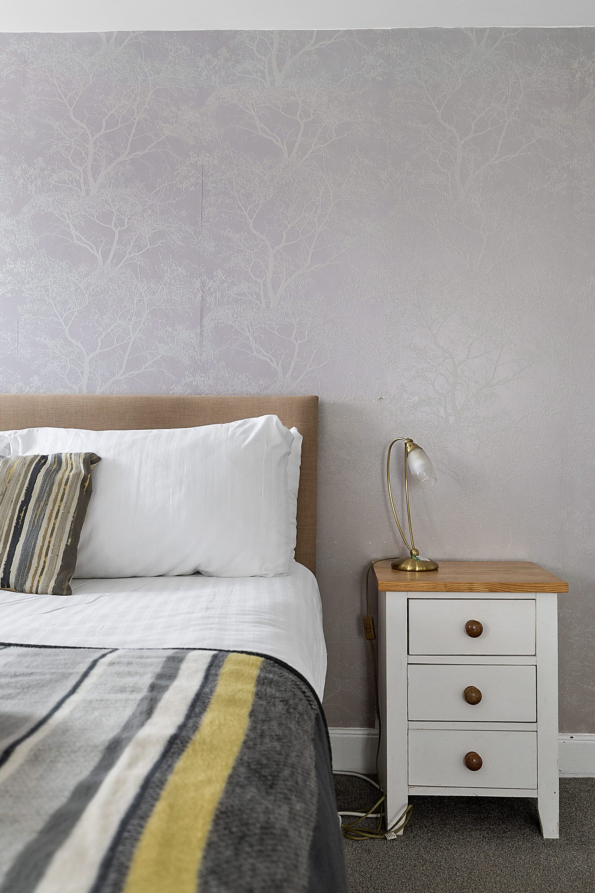 A neatly made bed with a striped blanket and pillows, a bedside table with a lamp, and a wall with a tree pattern.