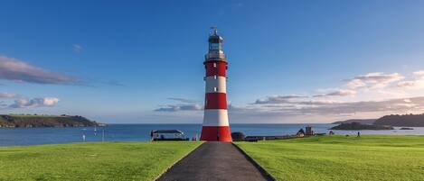A lighthouse with a red and white striped tower, a paved walkway leading to it, and a grassy area with a view of the sea and distant hills.