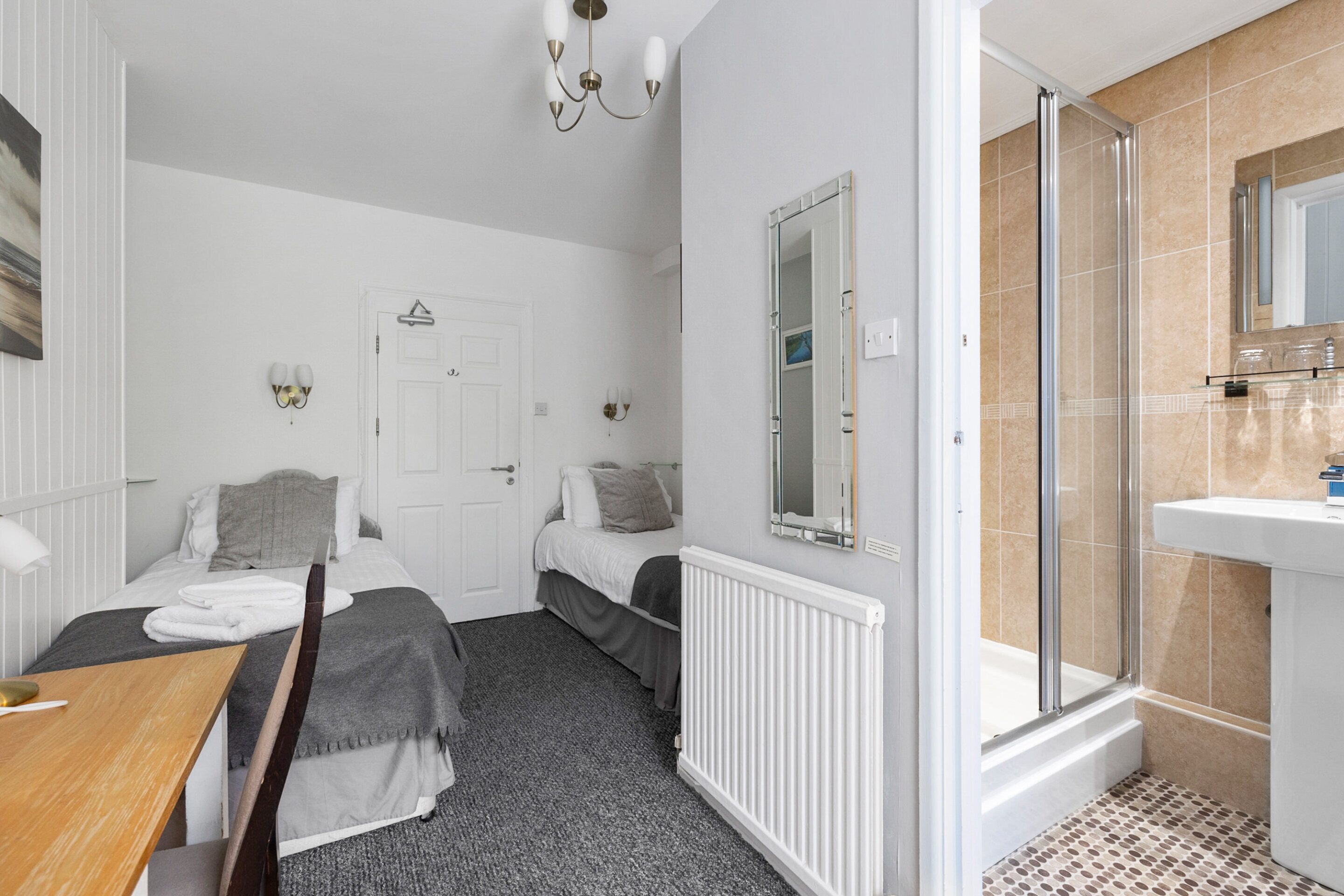A bedroom with two beds, a wooden desk, a radiator, and a bathroom with a shower and sink.