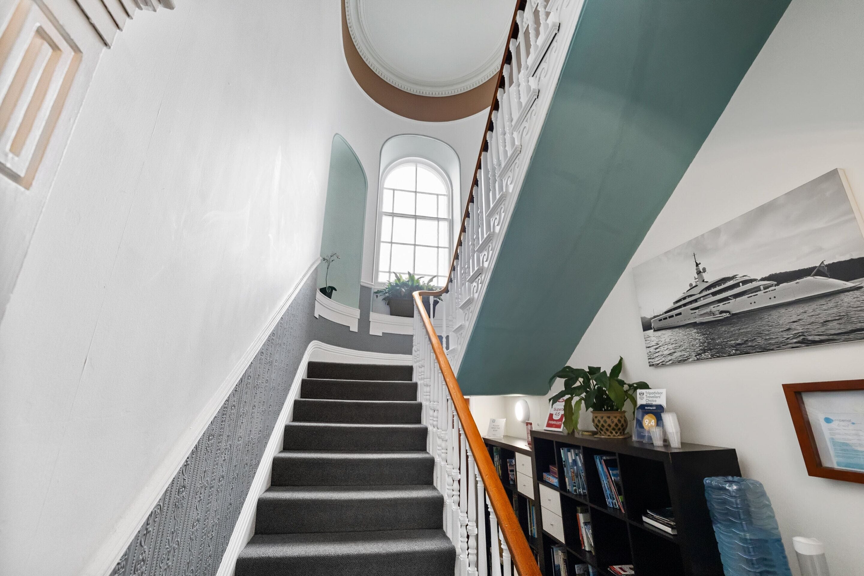 A staircase with a wooden handrail, a bookshelf with books, a framed picture of a ship, and a potted plant.