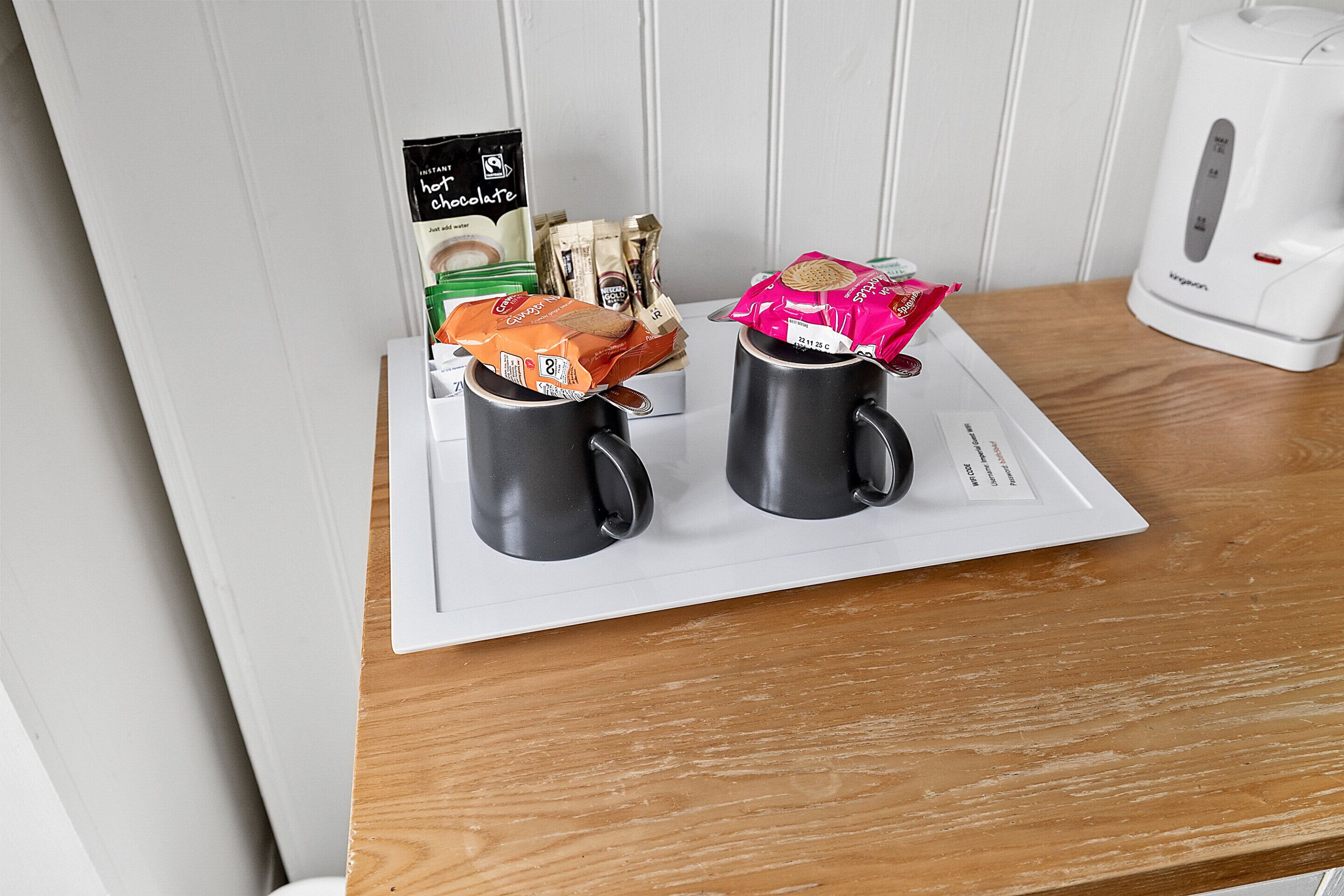 A table with two black mugs, a packet of instant coffee, a packet of tea, and a packet of biscuits.