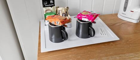 A table with two black mugs, a packet of instant coffee, a packet of tea, and a packet of biscuits.