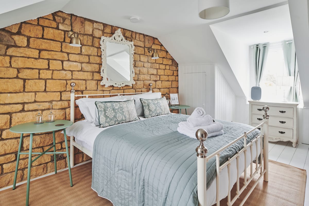 A bedroom with a brick wall, a bed with a blue quilt, a small round table, and a dresser.