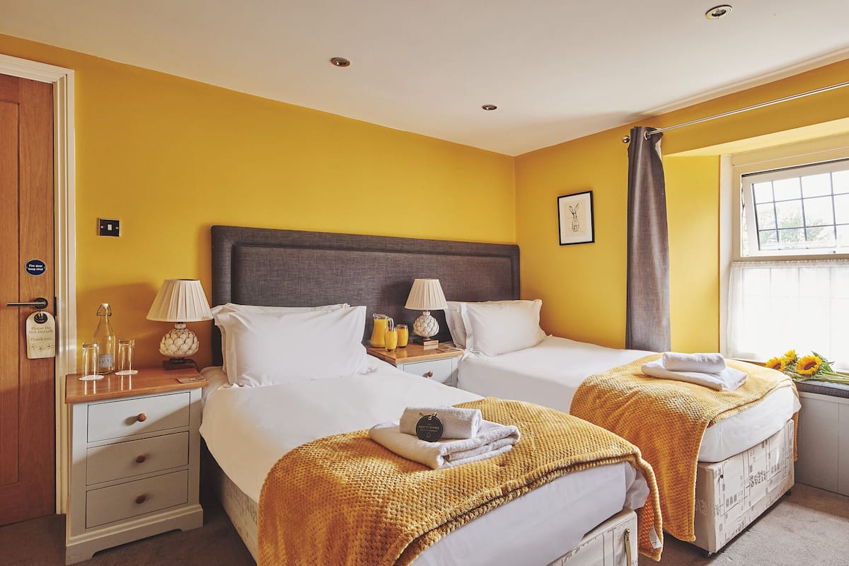 A hotel room with two beds, a wooden nightstand with a lamp, a small table with a lamp, and a window with a view of the outdoors.