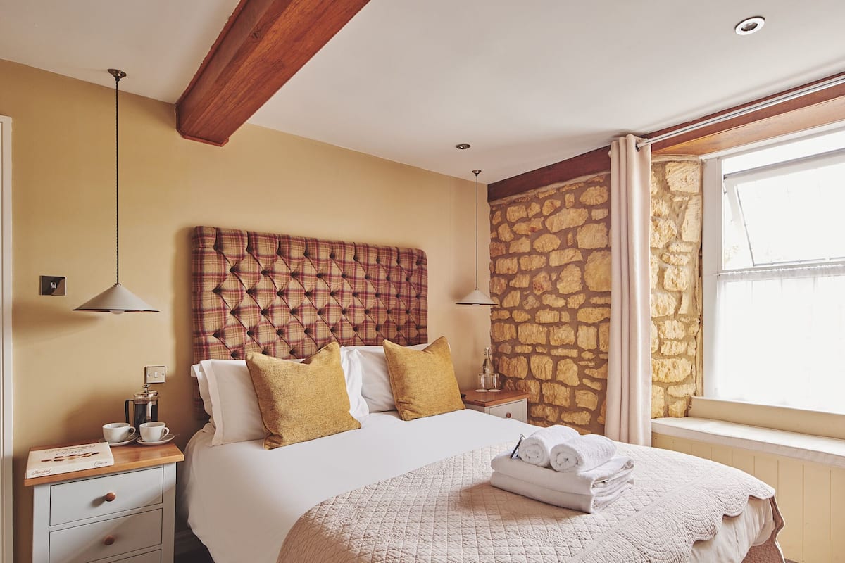 A neatly arranged bedroom with a large bed, a bedside table with a lamp, a stone wall, and a window with curtains.
