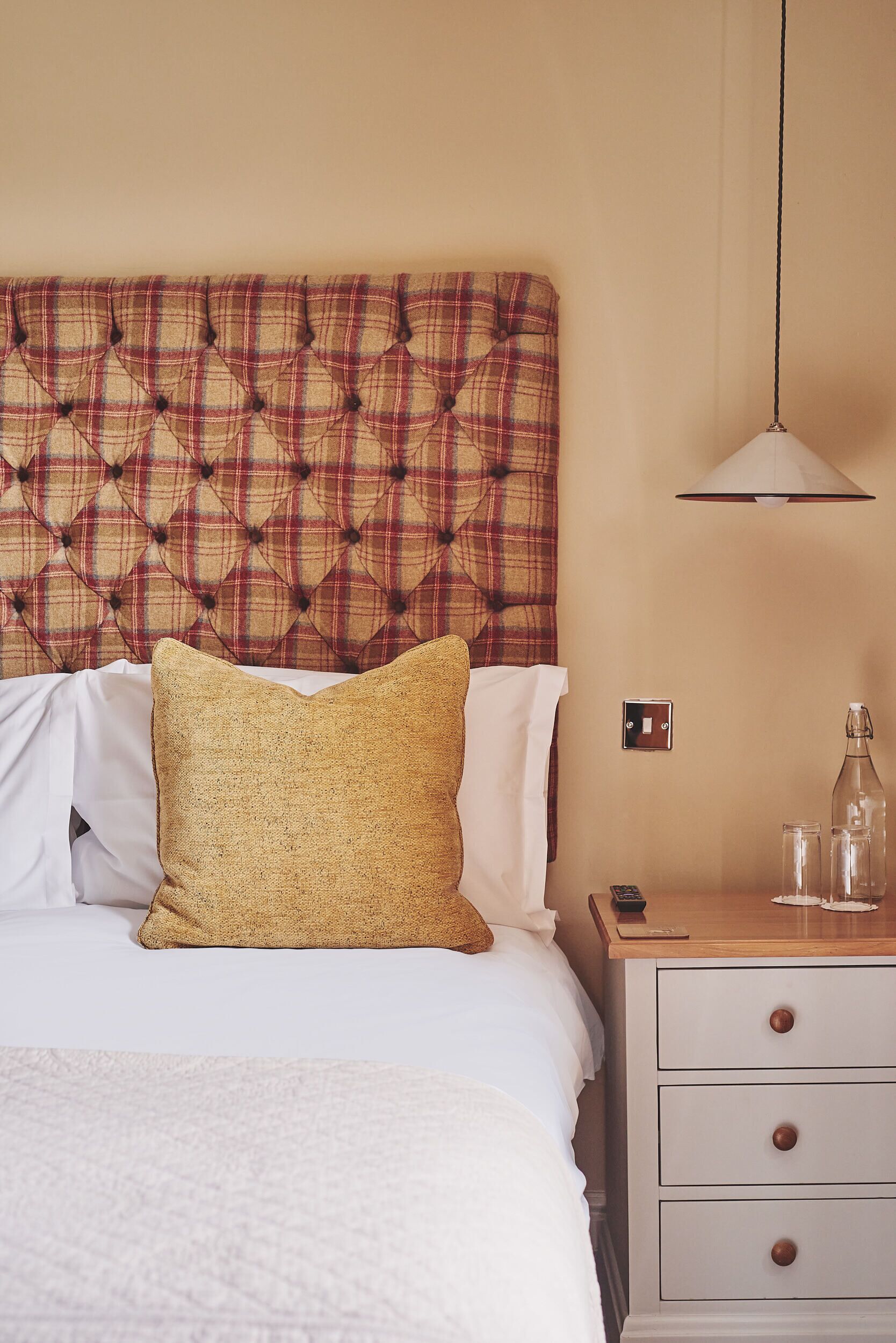 A neatly made bed with a plaid headboard, a bedside table with a lamp, and a small drawer unit.