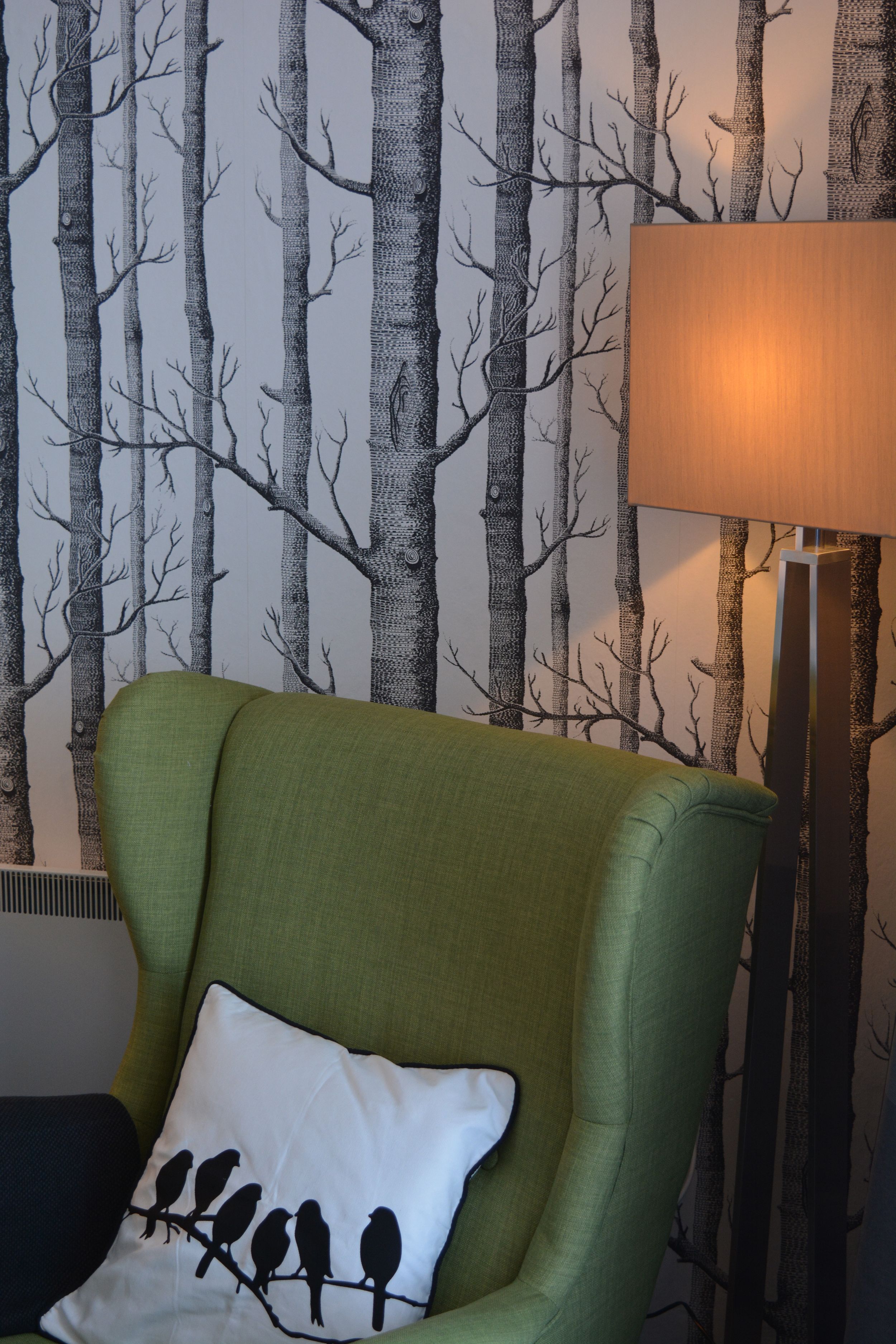 A green armchair with a white pillow featuring bird silhouettes, positioned in front of a wall with a black and white tree pattern.