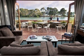 A living room with a view of a lake, outdoor seating, and a table with books and a cup. - The Waterfront Wynyard (Wynyard)