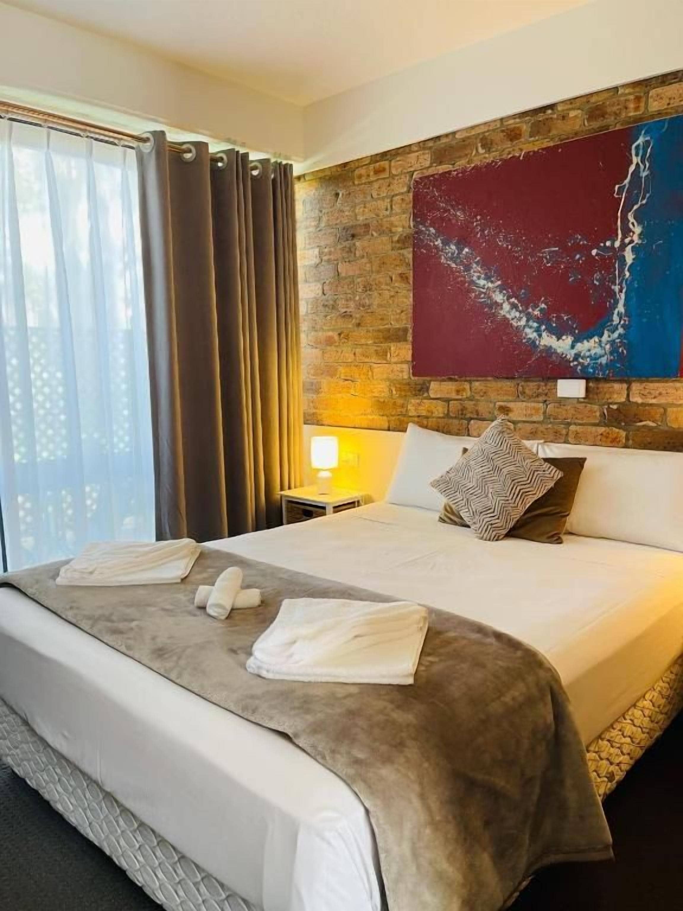 Habitación de hotel con una cama grande, una pared de ladrillo, un cuadro y una ventana con cortinas.
