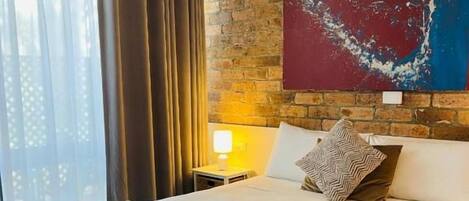 Habitación de hotel con una cama grande, una pared de ladrillo, un cuadro y una ventana con cortinas.
