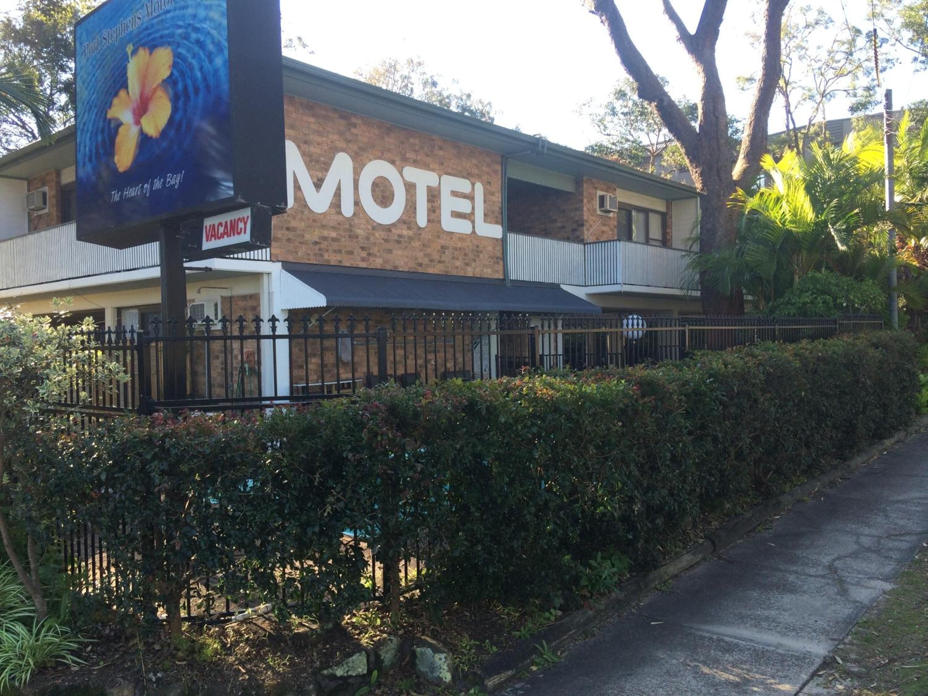 A motel with a large sign, a fenced parking area, and a pathway leading to the entrance.