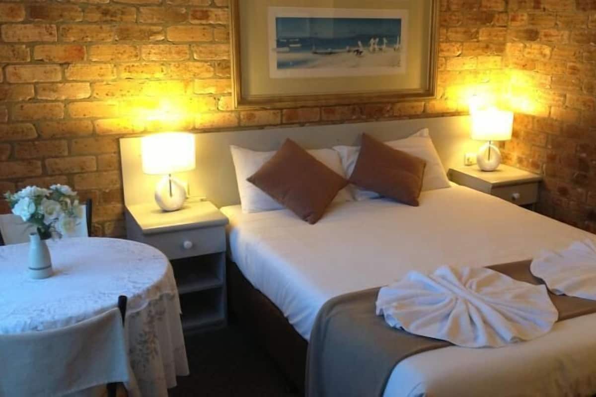 A hotel room with a brick wall, a bed with white bedding and brown pillows, two bedside lamps, a small round table with a vase of flowers, and a framed picture on the wall.