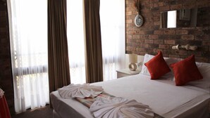 Une chambre d’hôtel avec un mur en briques, un lit avec du linge de lit blanc et des oreillers rouges, une table de chevet avec une lampe, et une grande fenêtre avec des rideaux.