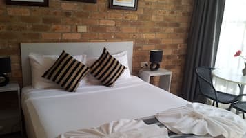 A hotel room with a brick wall, a bed with white bedding, striped pillows, a bedside table with a lamp, a chair, and a table with a vase of flowers.