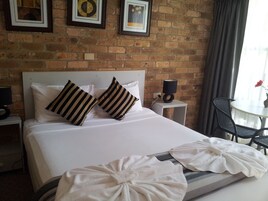 A hotel room with a brick wall, a bed with white bedding, striped pillows, a bedside table with a lamp, a chair, and a table with a vase of flowers.