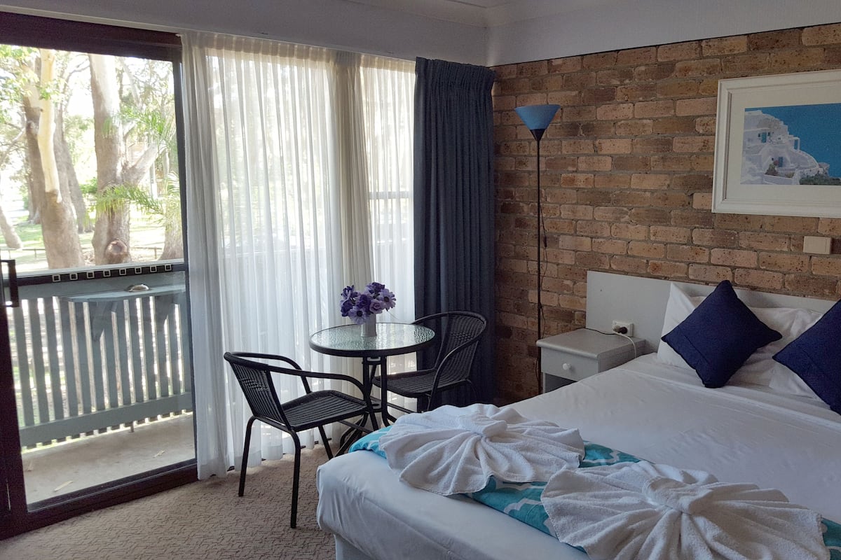 A hotel room with a bed, a desk, a chair, a window with curtains, and a brick wall.
