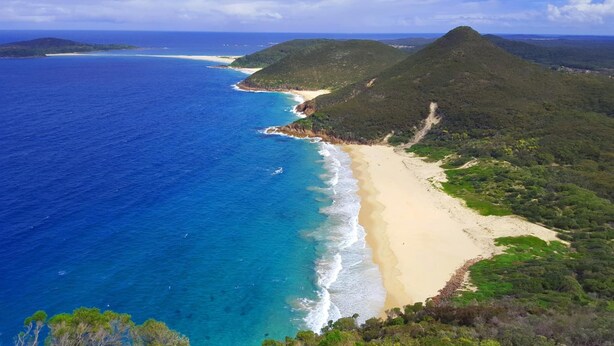 Un paysage côtier avec une plage, une eau d’un bleu limpide et un relief vallonné.