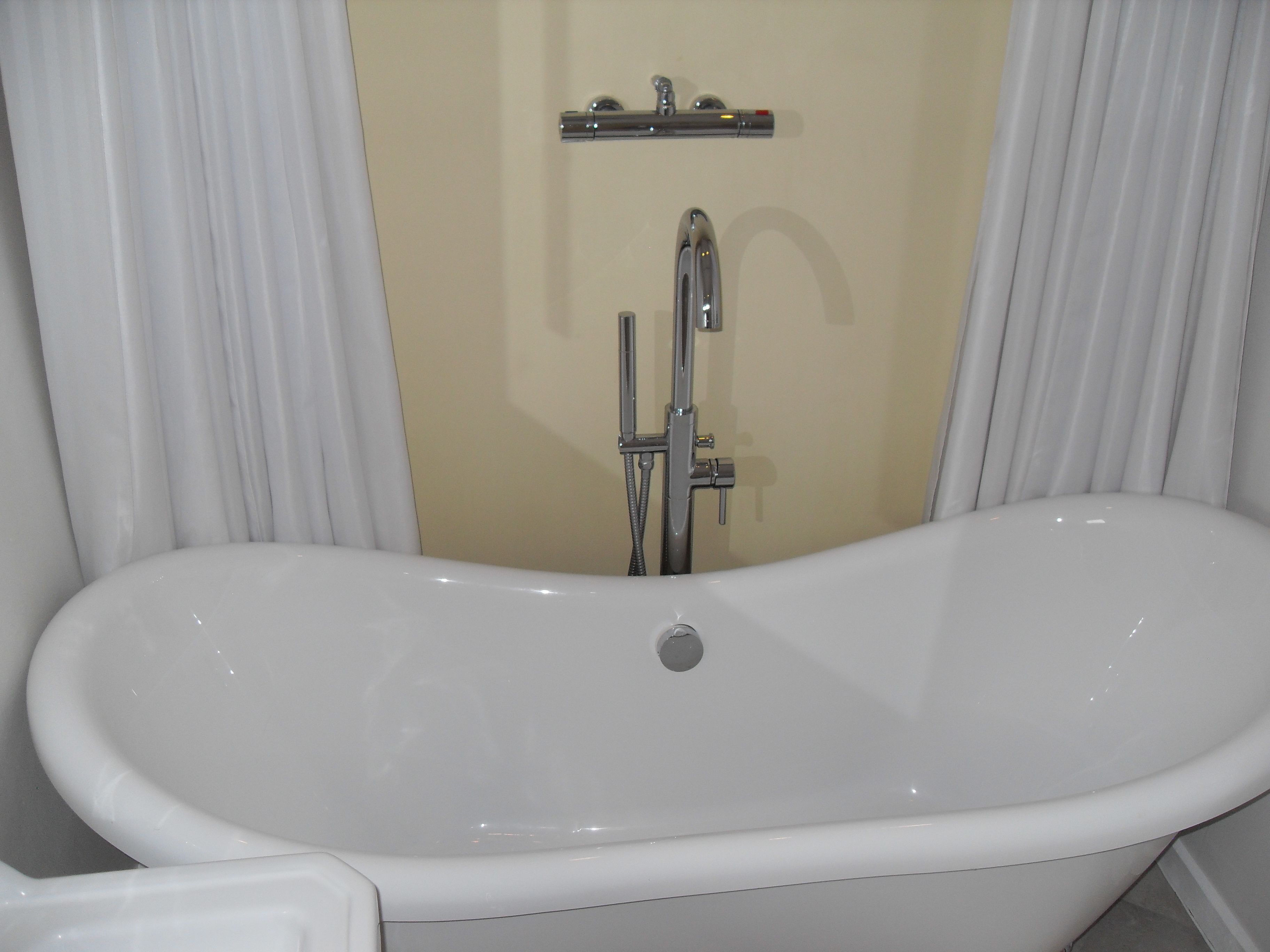 A white freestanding bathtub in a bathroom with beige walls and white curtains.
