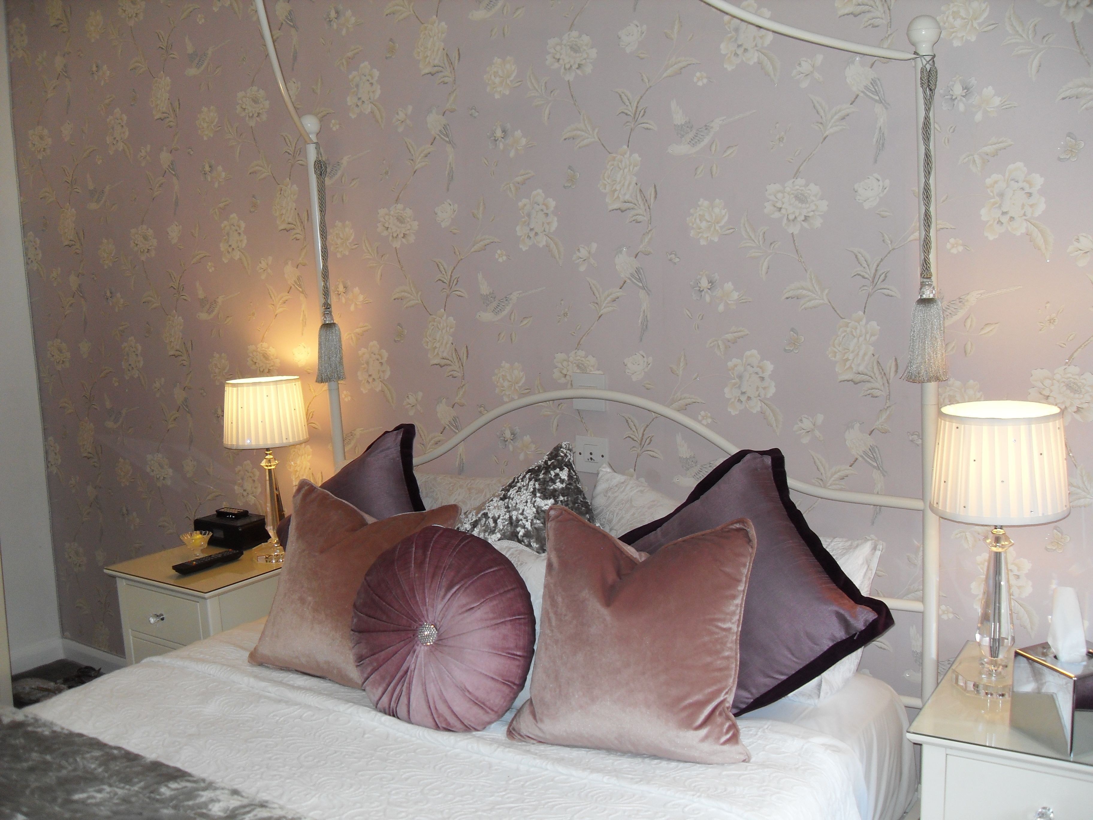 A neatly made bed with a white comforter and decorative pillows, a nightstand with a lamp, and a wall with floral wallpaper.