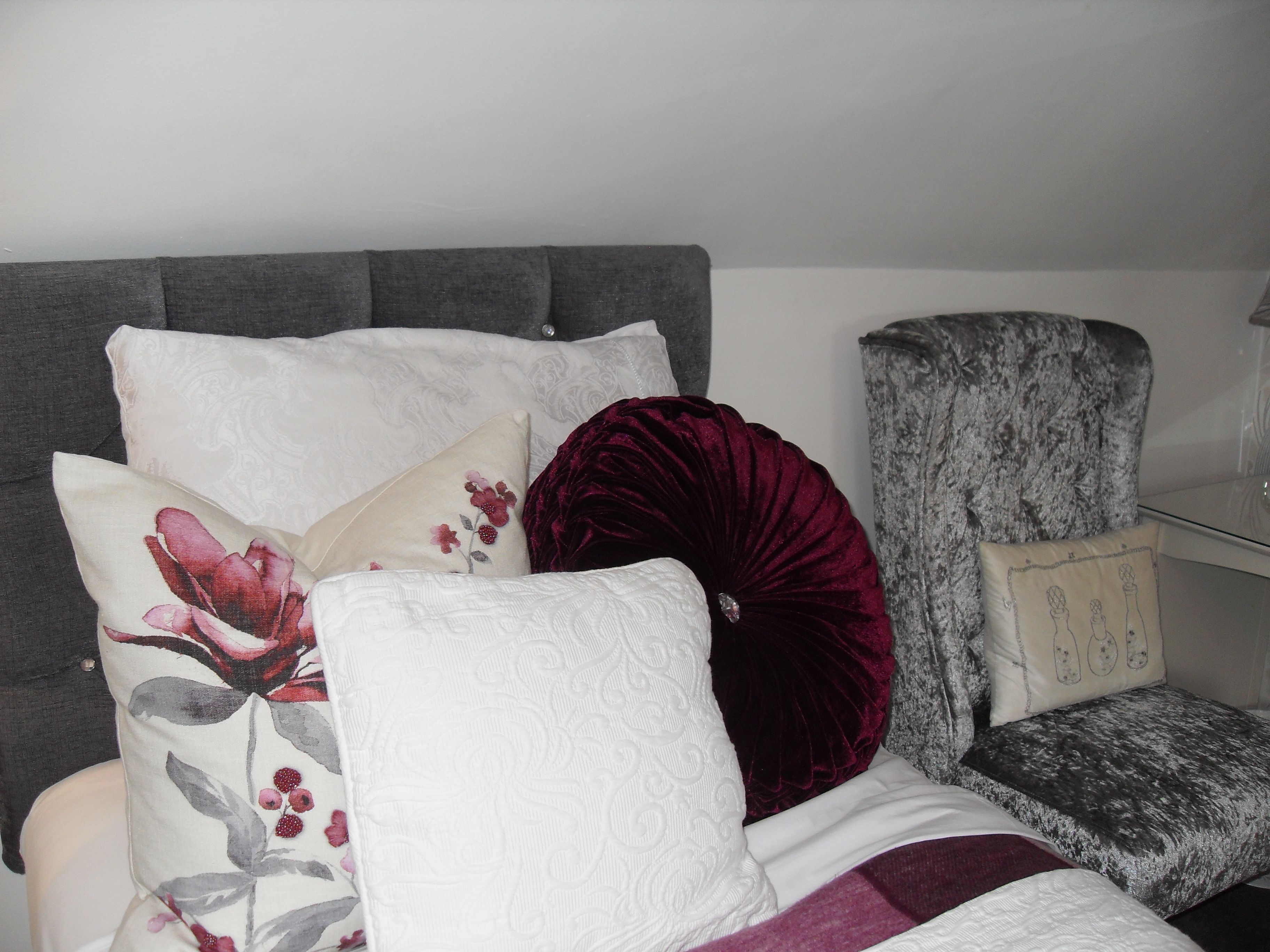 Un dormitorio con un cabecero tapizado en gris, un sillón tapizado en blanco y una cama con múltiples almohadas, incluyendo una roja.