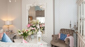 A well-lit room with a patterned wallpaper, a sofa with cushions, a chair, a table with a vase of flowers, and a mirror with an ornate frame. - Jessop House (Tewkesbury)