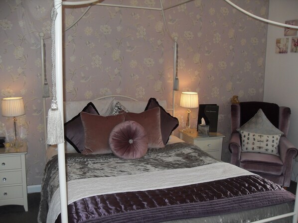 A four-poster bed with a canopy, a nightstand with a lamp, a chair with a cushion, and a wall with floral wallpaper. - Jessop House (Tewkesbury)