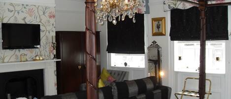A four-poster bed with a floral bedspread, a dark wooden four-poster canopy, a chandelier, a fireplace, a television, and a window with a view of the street.