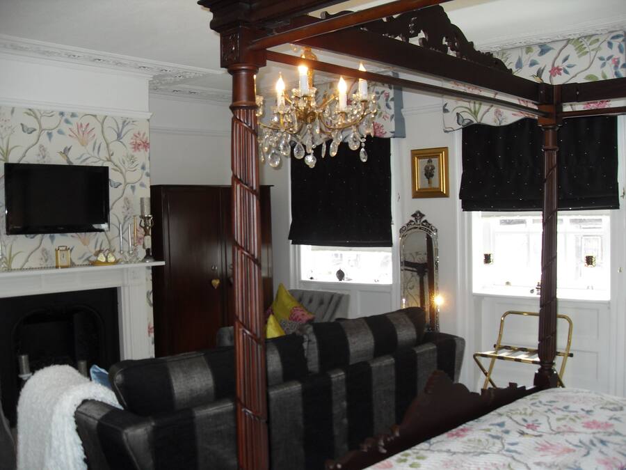 A four-poster bed with a floral bedspread, a dark wooden four-poster canopy, a chandelier, a fireplace, a television, and a window with a view of the street.
