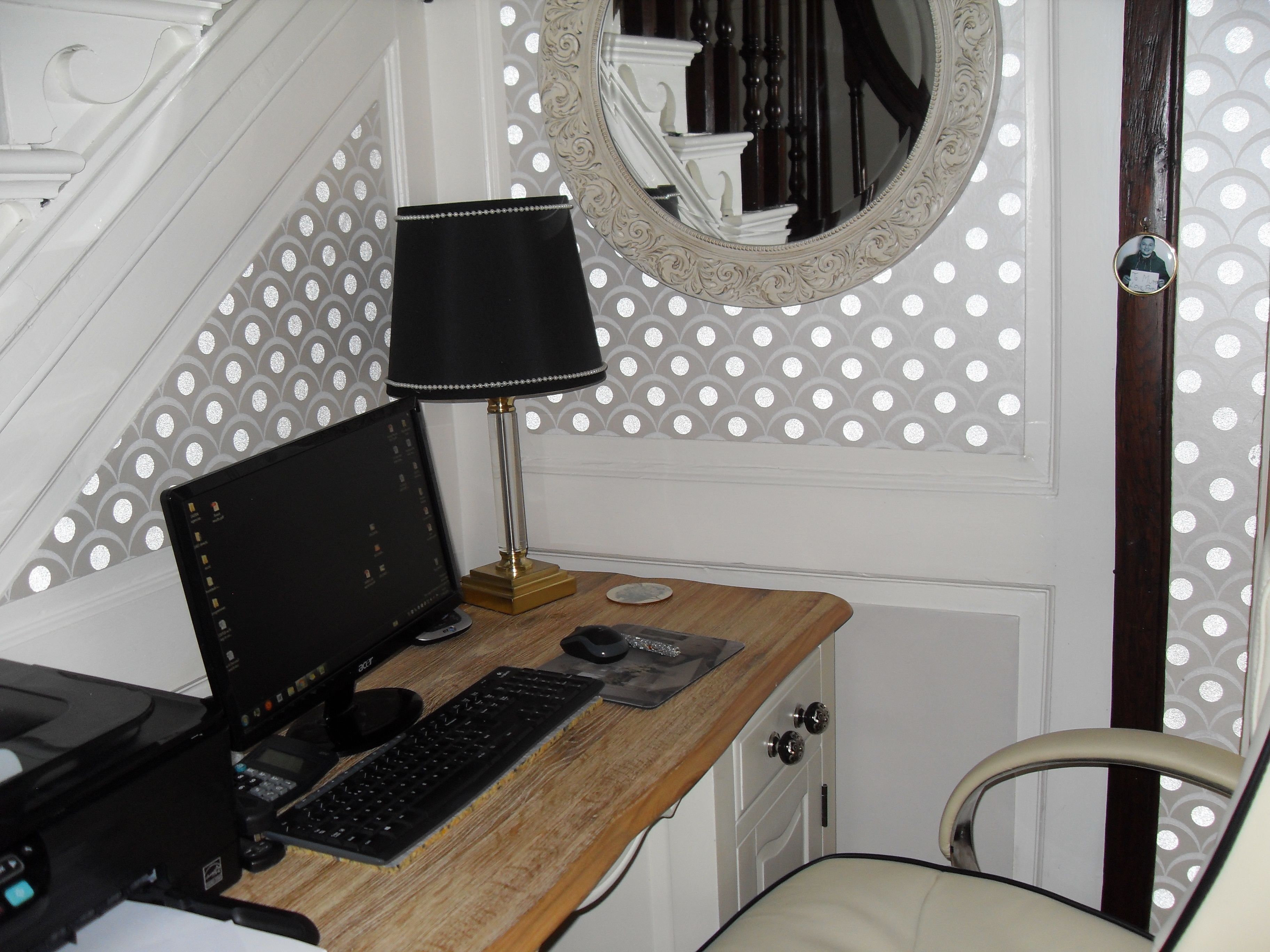 Een computerbureau met een monitor, toetsenbord en lamp in een kamer met behang met een patroon en een spiegel.