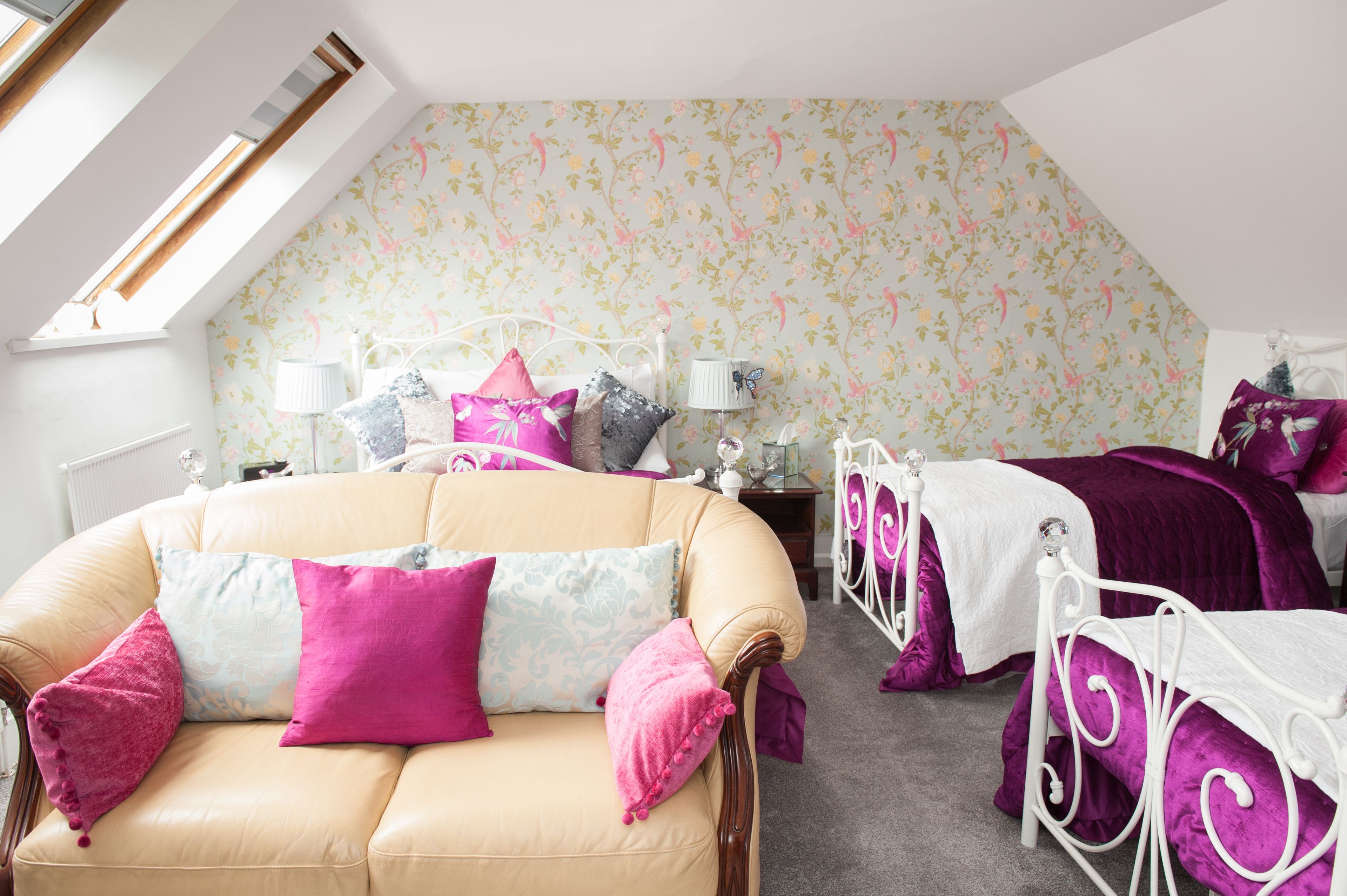 Ein Schlafzimmer mit geneigter Decke, blumigem Wandpapier, einem beigefarbenen Sofa mit rosa und blauen Kissen, einem weißen Metallbettgestell und einem weißen Nachttisch mit Lampe.