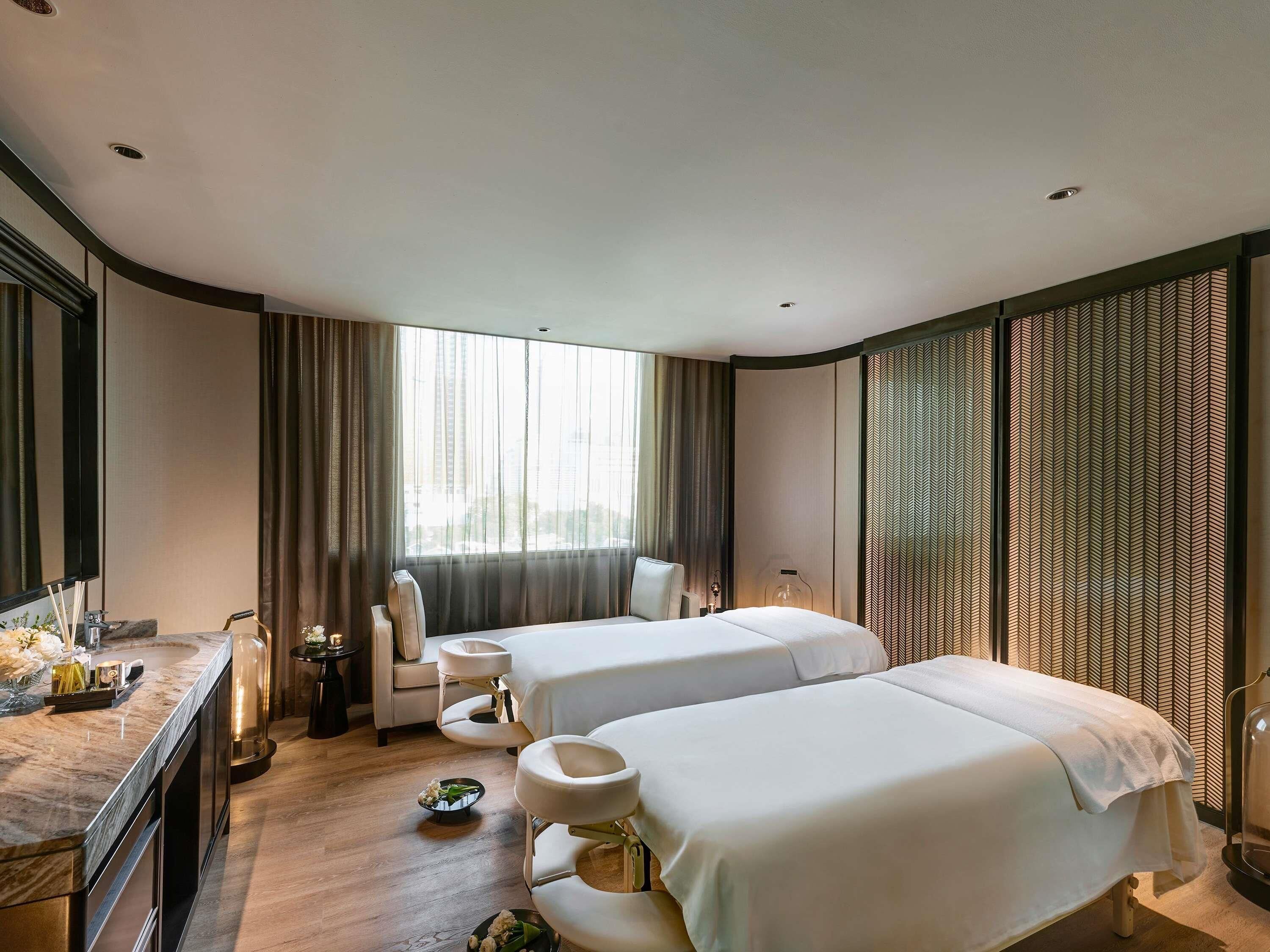 A hotel room with two beds, a sitting area with a chair, a vanity with a mirror, and a large window with curtains.
