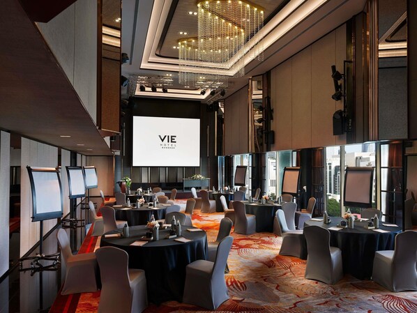 Un salón de conferencias en el VIE HOTEL BANGKOK con múltiples mesas, sillas, una pantalla grande y un candelabro.