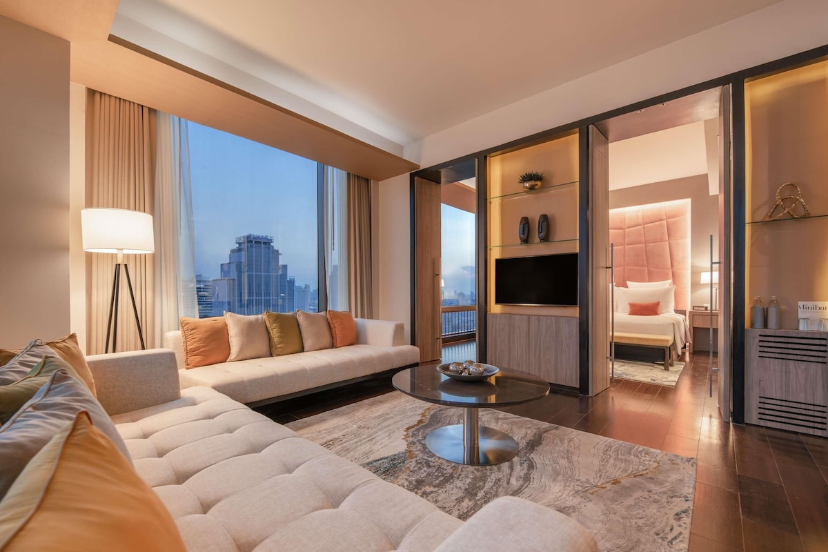 A modern hotel room with a large sofa, a coffee table, and a TV. The room has a city view and a balcony.
