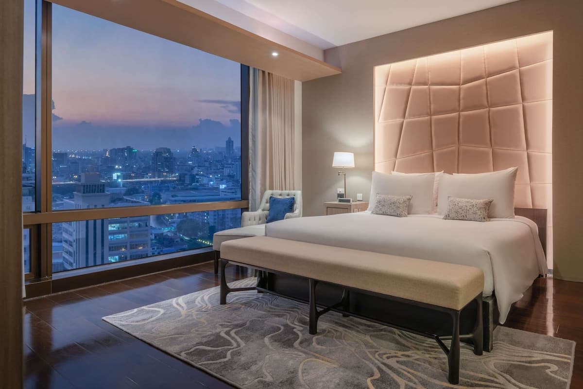 Ein modernes Hotelzimmer mit einem großen Bett, einer Bank, einem Nachttisch mit Lampe und Blick auf eine Stadtlandschaft durch große Fenster.