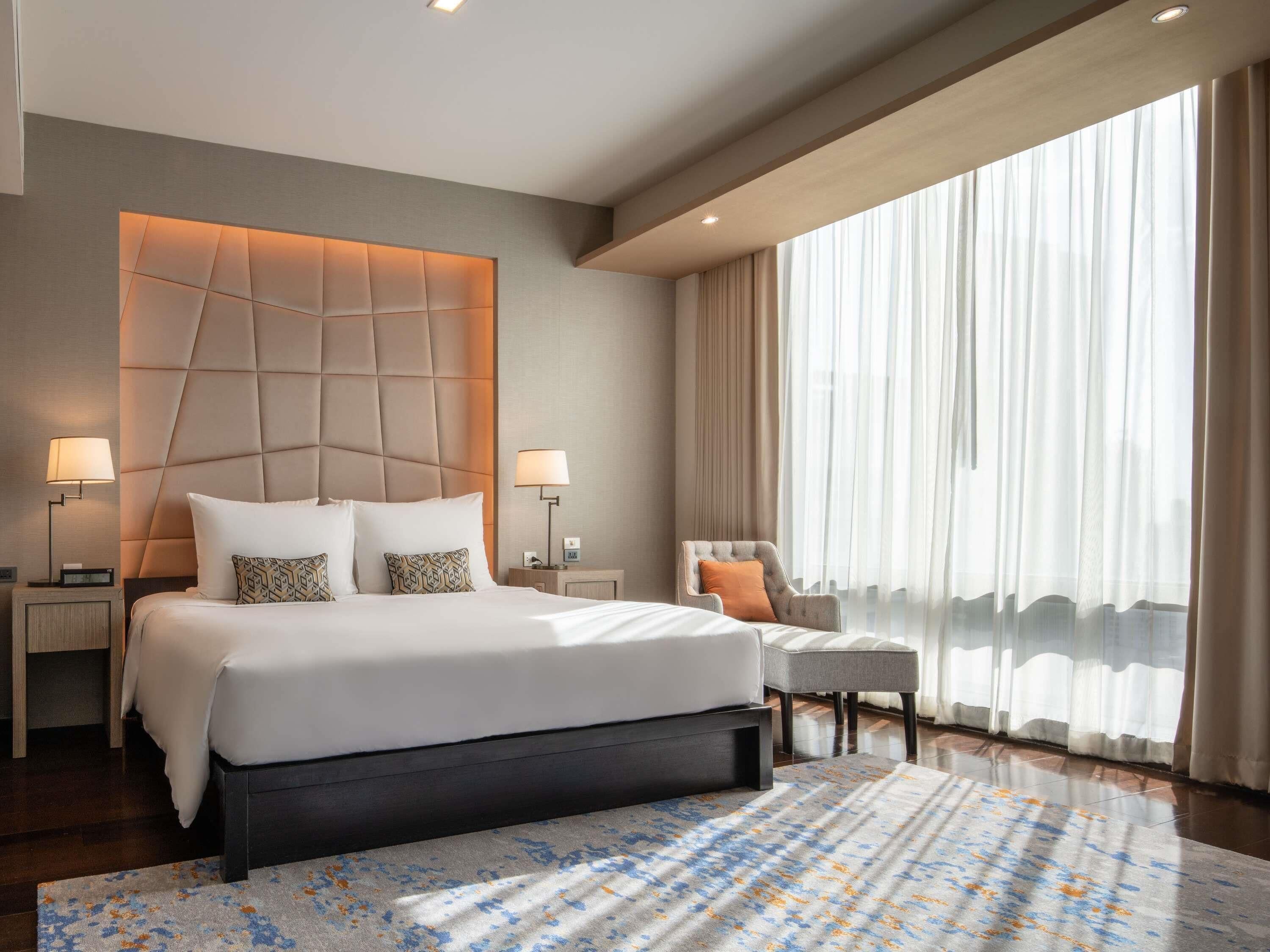 A modern hotel room with a large bed, two bedside tables, a chair, a large window with sheer curtains, and a patterned rug.
