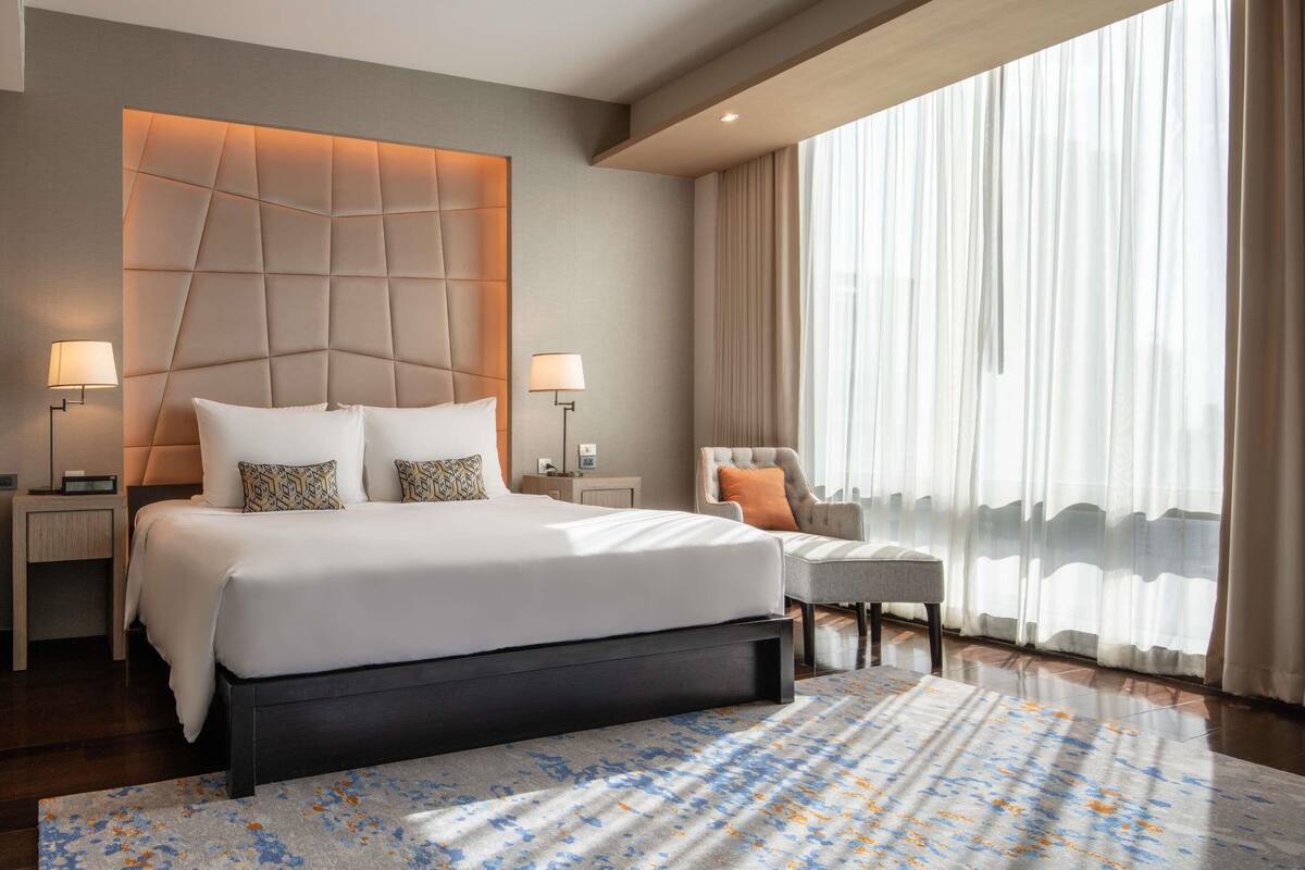 Une chambre d’hôtel moderne avec un grand lit, deux tables de chevet, une chaise, une grande fenêtre avec des rideaux légers et un tapis à motifs.