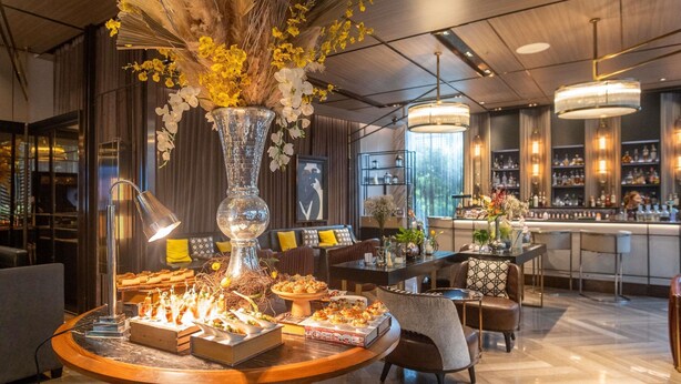 Een moderne bar met een ronde houten tafel waarop hapjes staan, een decoratieve vaas met bloemen en een bar met flessen.