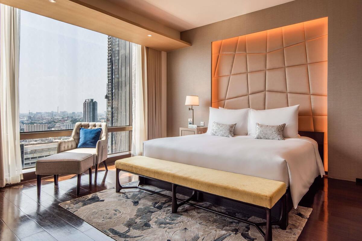 Ein Hotelzimmer mit einem großen Bett, einer Sitzbank, einem Nachttisch und Blick auf die Stadt.
