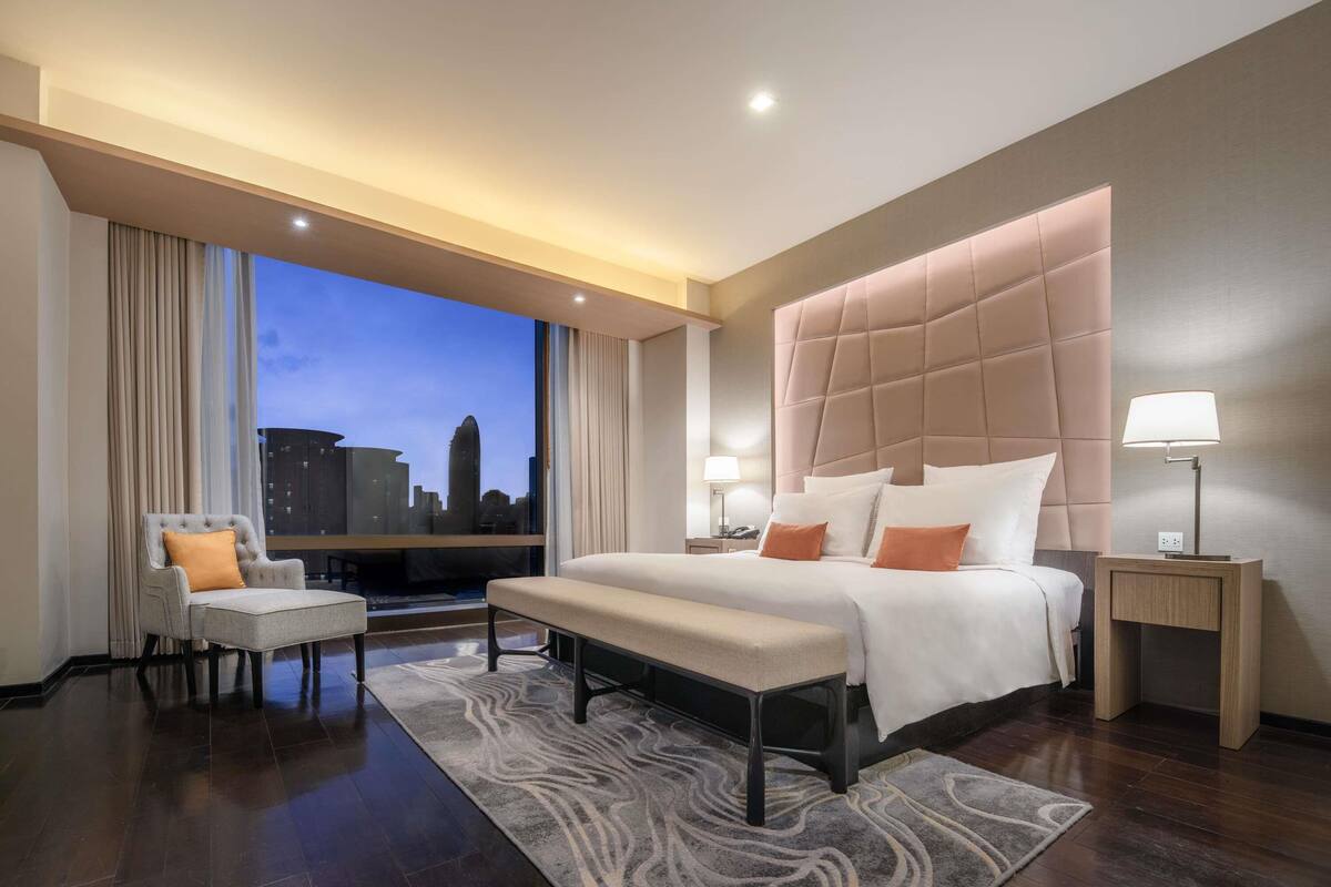 Une chambre d’hôtel moderne, dotée d’un grand lit, d’une banquette, d’une chaise, d’une table de chevet et offrant une vue sur le paysage urbain.