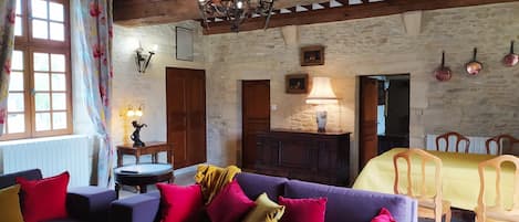 A rustic room with a wooden ceiling, a purple sofa with colorful cushions, a dining table with chairs, and a stone wall with artwork.
