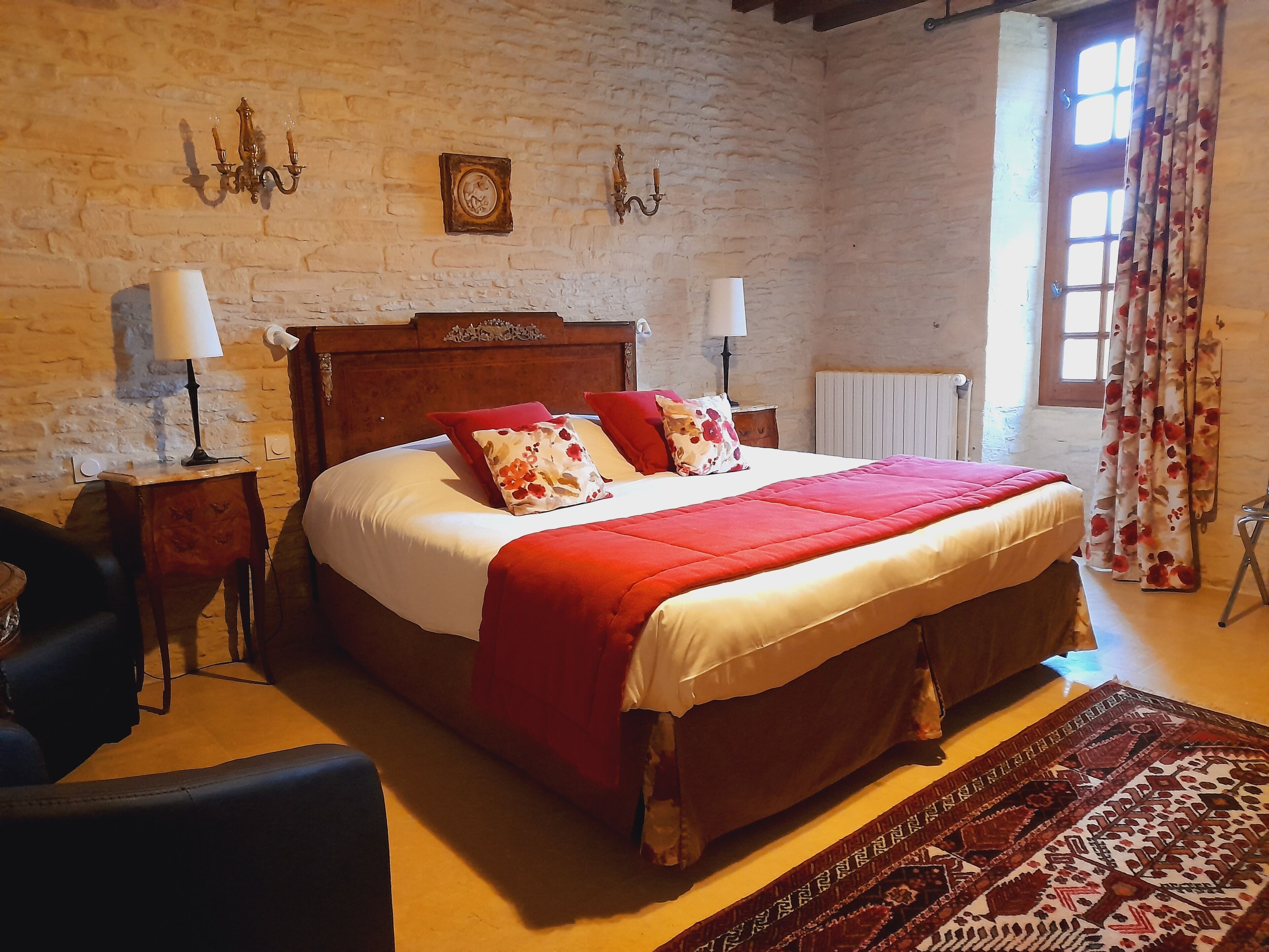 A traditional room with a brick wall, a wooden bed with red and white bedding, two bedside tables with lamps, a chair, and a patterned rug.