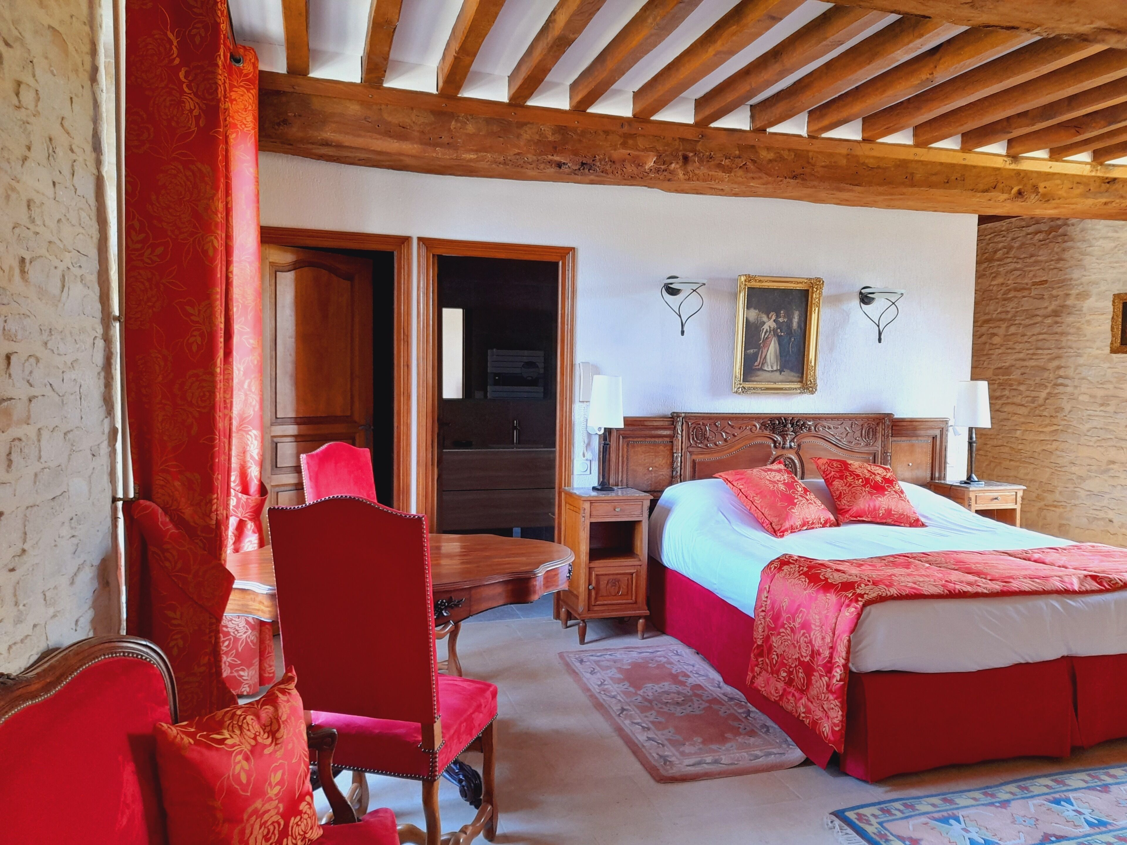 A bedroom with a wooden ceiling, a bed with red bedding, a wooden nightstand, a chair, and a door leading to another room.