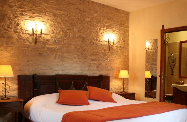A hotel room with a stone wall, a wooden bed with two orange pillows, two bedside lamps, and a mirror above the bed. - Hotel Manoir de Mathan (Crepon)